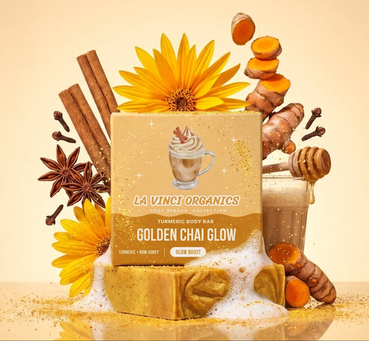 A boxed tea product labeled 'La Vinci Organics Golden Chai Glow' stands on a reflective surface with cinnamon sticks, star anise, turmeric root, honey, sugar, and yellow flowers arranged around it against a warm beige background.